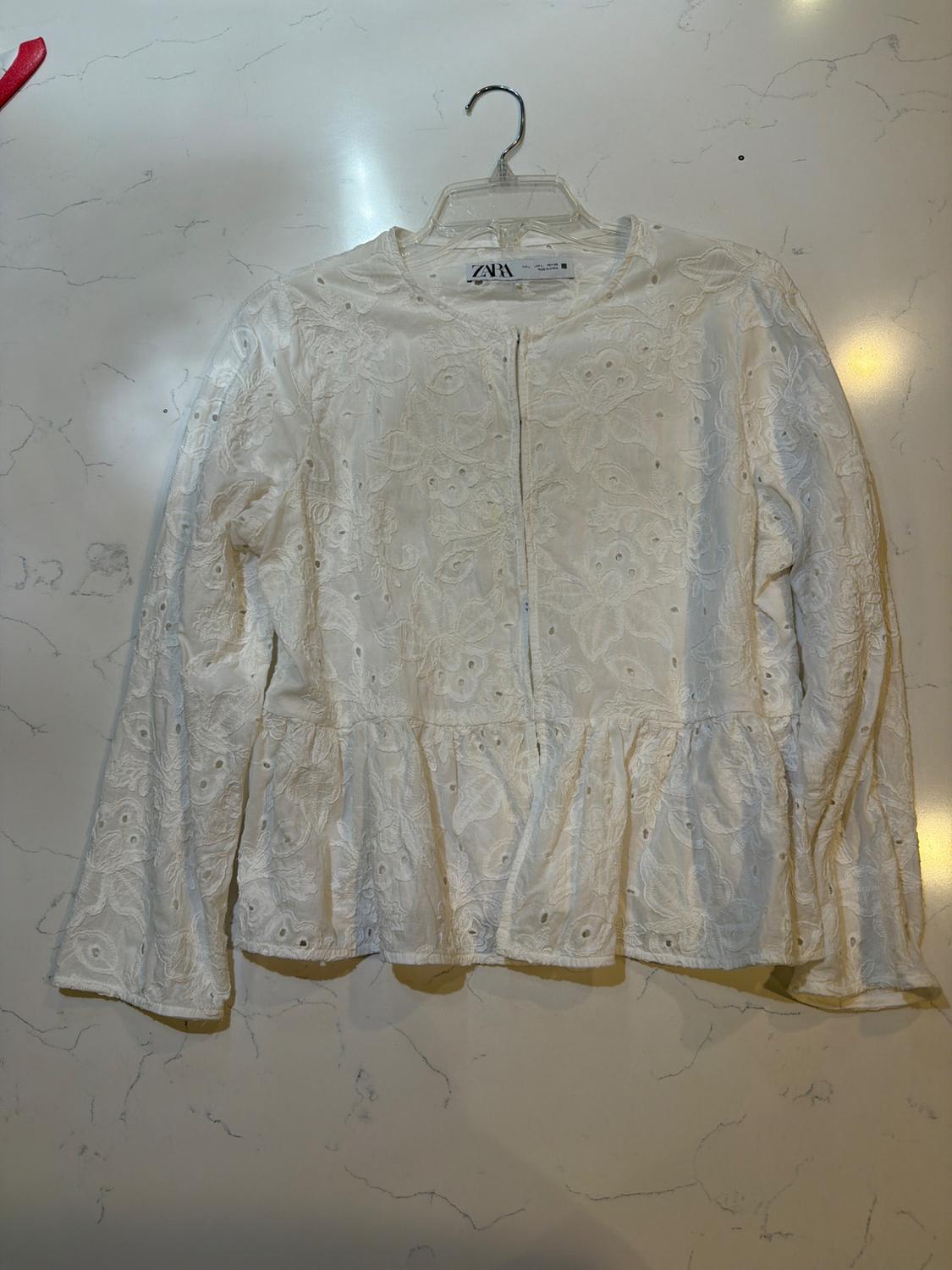 White long-sleeved blouse with eyelet embroidery and a ruffle, on a hanger on a light marble surface. Visible text: ZARA.
