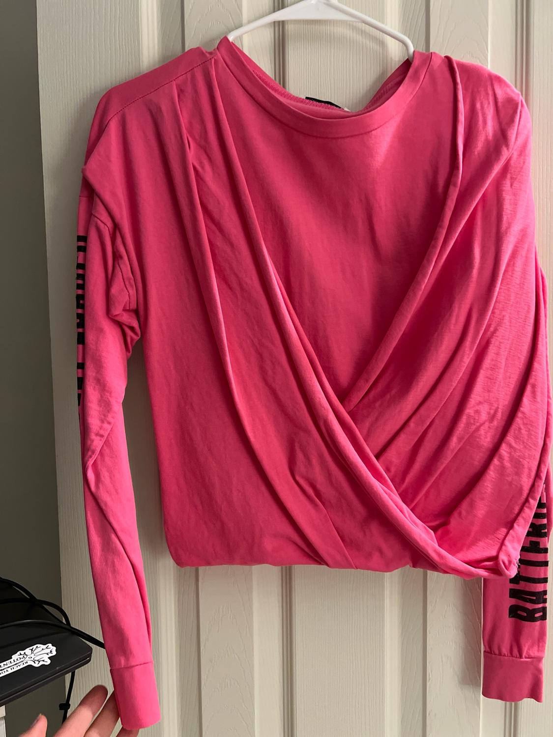 Pink long-sleeve shirt with a crossed design hanging on a white door. Sleeves with black text. It reads REACH YOUR POTENTIAL.
