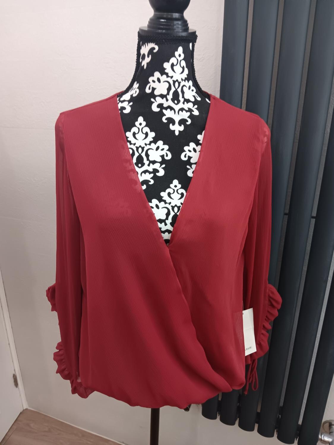 Red, long-sleeved wrap blouse with ruffles on a black and white patterned mannequin. Label with the text BASIC.