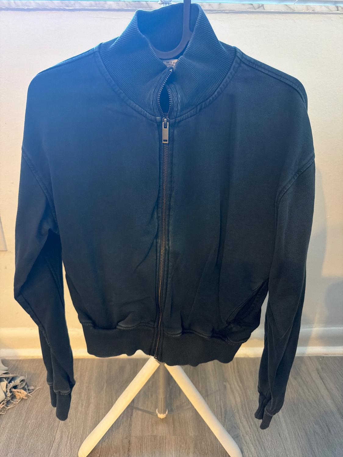 Casual black jacket with a front zipper and ribbed high neck, hanging on a white tripod on a wooden floor.