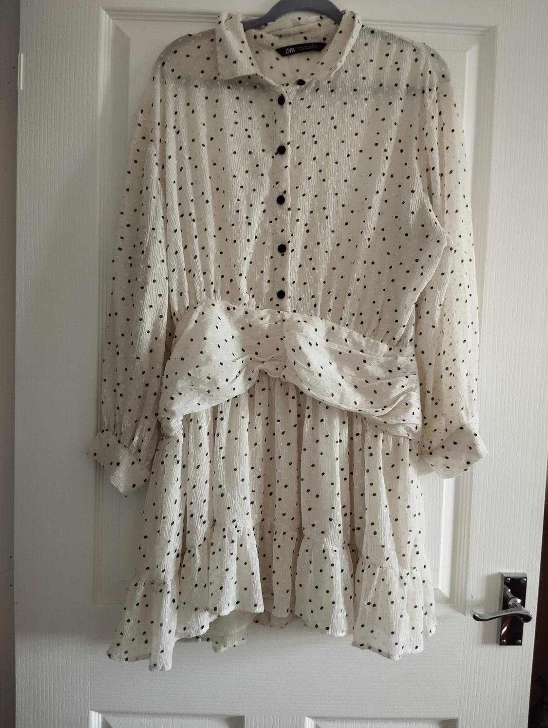 White polka-dot dress hanging on a door. ZARA label. Long sleeves, front buttons, draped waist and ruffled hem.
