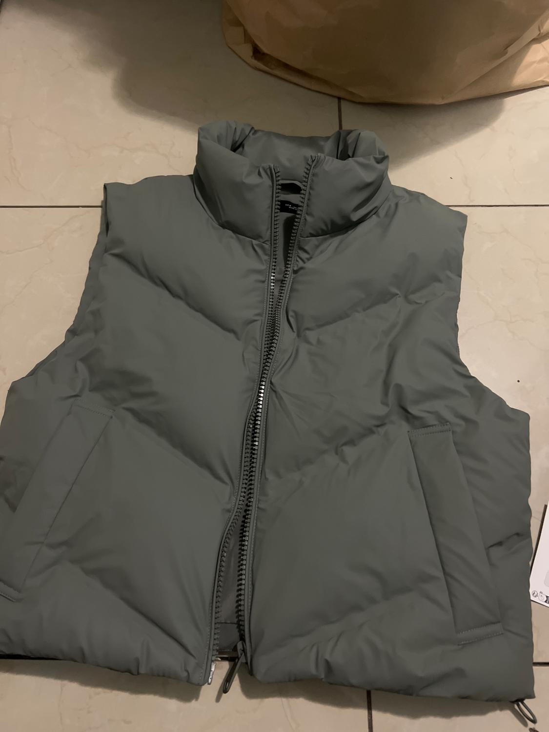 Grey padded gilet with front zip and high neck, placed on a light tiled floor.
