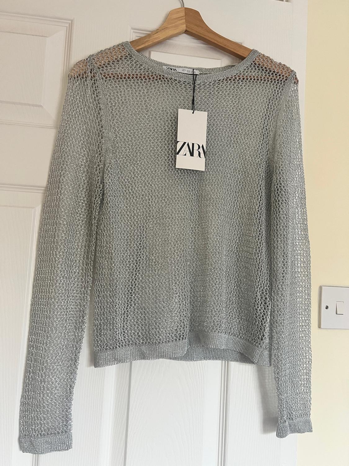 Grey open-knit jumper hanging on a wooden hanger, with a visible ZARA label, on a white door.
