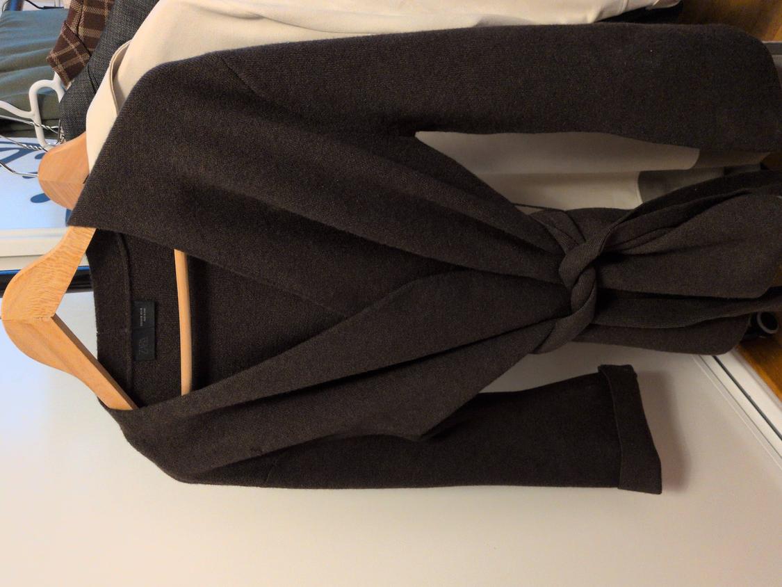 Dark brown coat with a knotted belt, hanging on a wooden hanger, against a background of light, patterned clothing.
