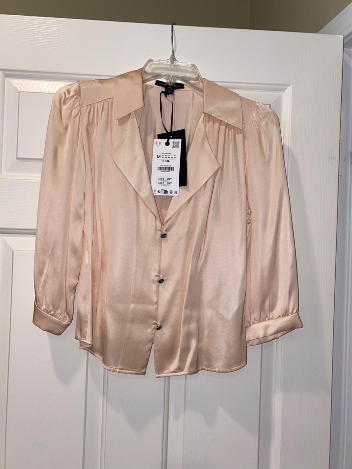 Peach-coloured satin blouse with front buttons, wide collar and puffed sleeves, hanging on a hanger in front of a white door.