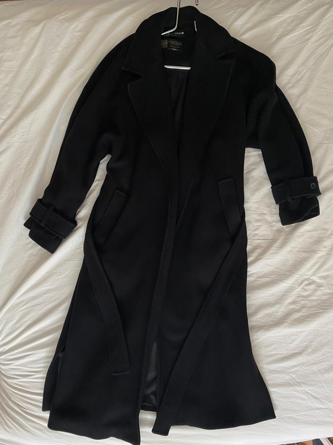 Long black coat with belt on hanger over crumpled white sheets. Relaxed atmosphere. Label with text MANTECO.