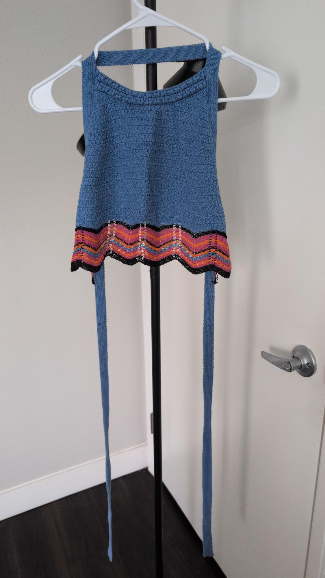 Blue knit top hanging on a white hanger. It features a wavy hem with pink, orange, and black stripes, and two long blue straps.