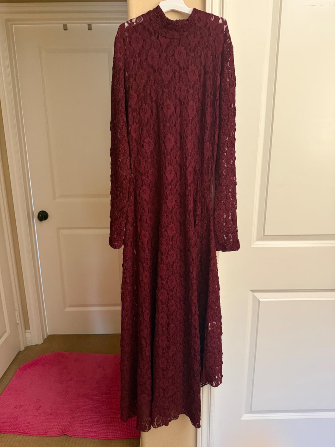 Long burgundy lace dress hanging on a white hanger, on a white door. Pink rug on the floor.