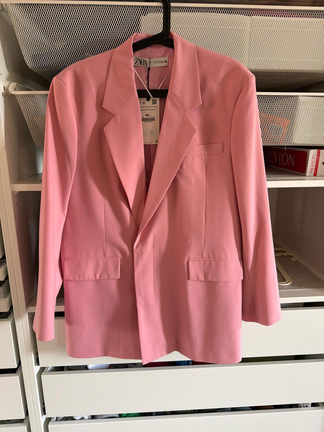 Pink blazer with lapels hanging in a white wardrobe. The ZARA label and a box with the text REVLON can be seen on a shelf.
