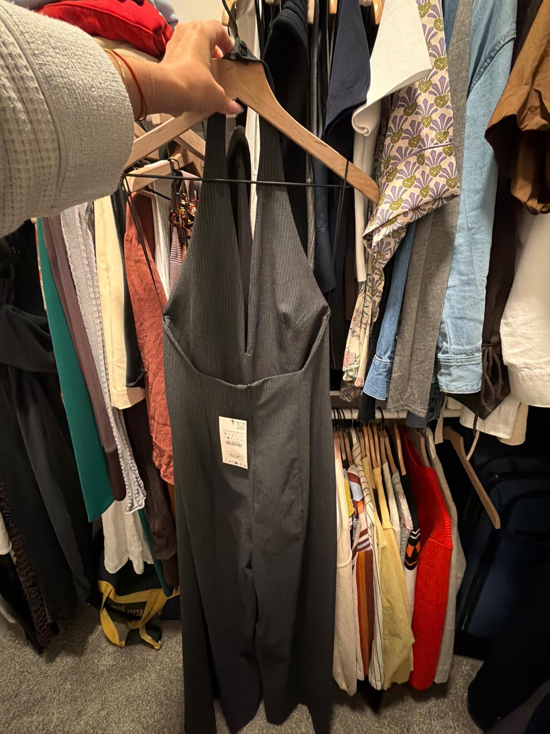 Person holding a gray jumpsuit on a rack full of varied clothing. Tag visible on the jumpsuit. Dressing room environment.