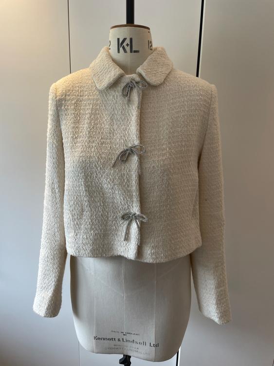 Rough-textured white jacket with bows on the front, displayed on a tailor's dummy against a neutral background.
