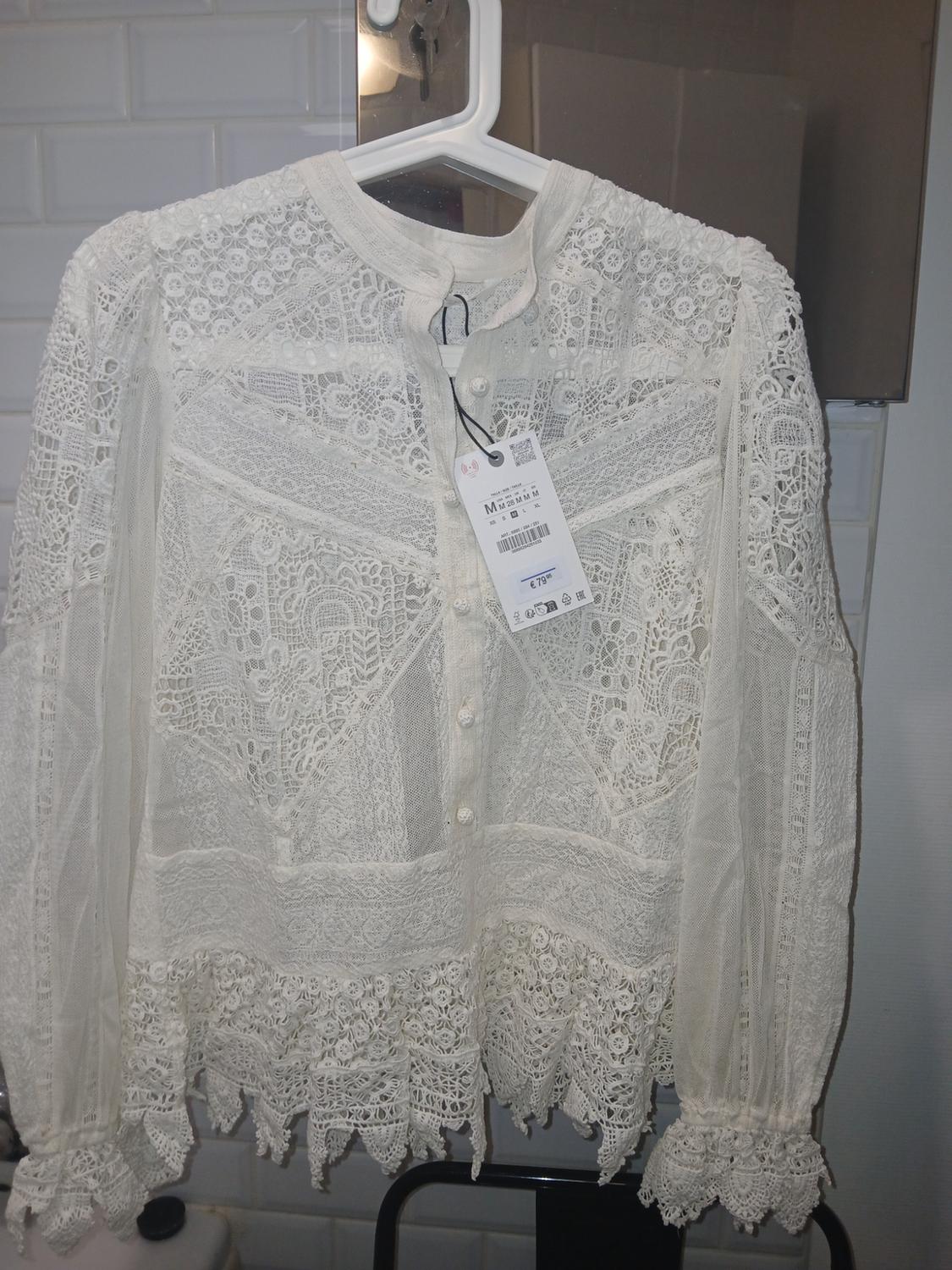 White lace blouse hanging on a hanger, with visible tag. White tiled background and mirror.