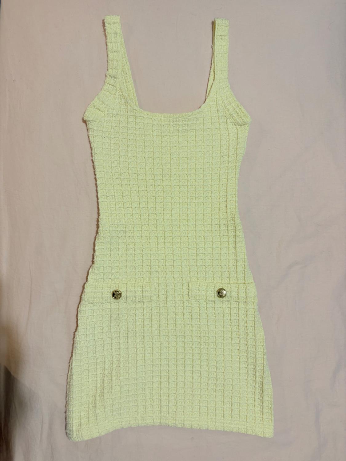 Light yellow short dress with straps, featuring a textured check fabric and two pockets with gold buttons, set against a light-coloured fabric background.