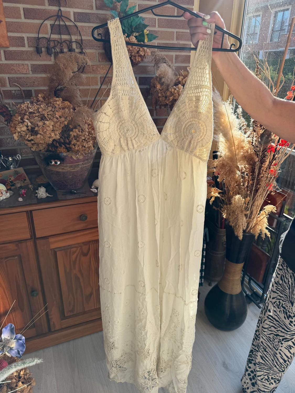 A white lace dress hangs on a hanger, held by a hand. The background shows a brick wall and a dried flower decoration.