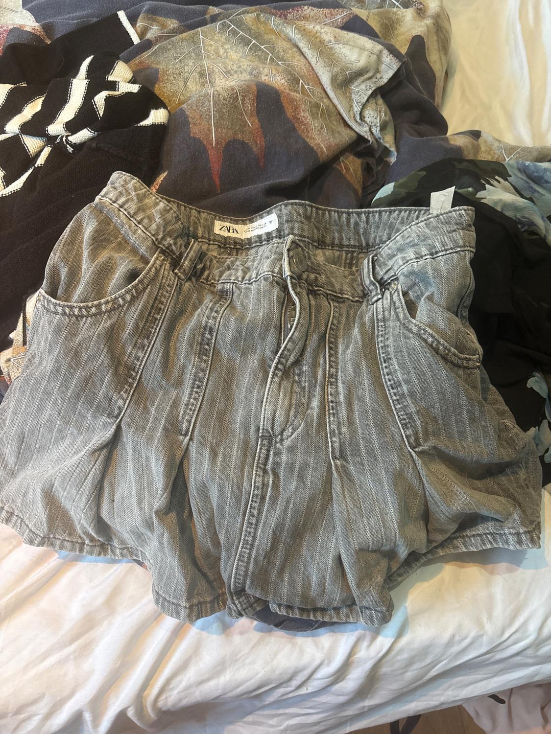 Grey striped shorts with pleats on an unmade bed with other garments. Label with the text ZARA. Casual and relaxed atmosphere.