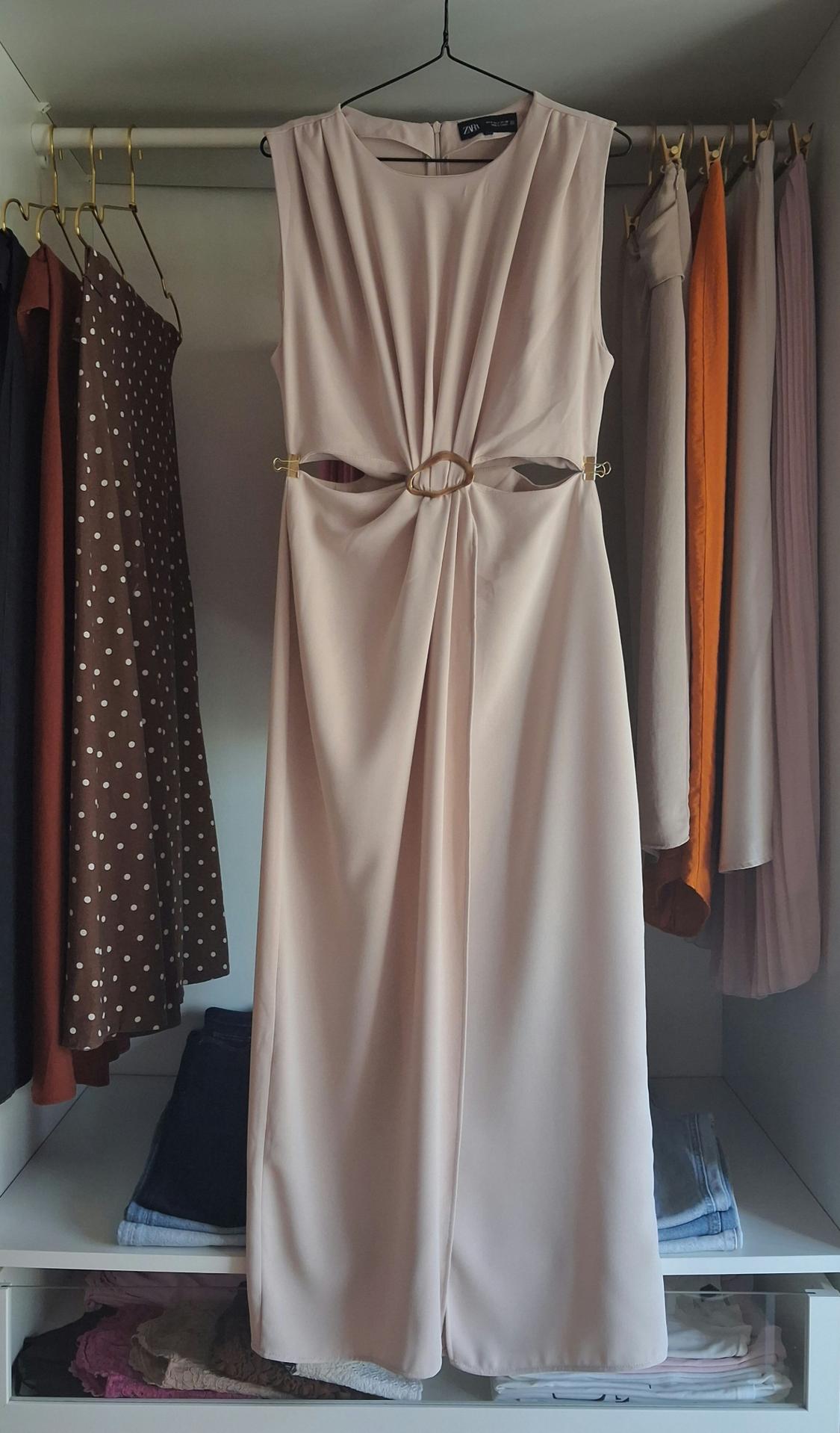 Beige sleeveless dress with cut-outs and a central ring, hanging in a wardrobe next to other garments. Text on the label: ZARA.
