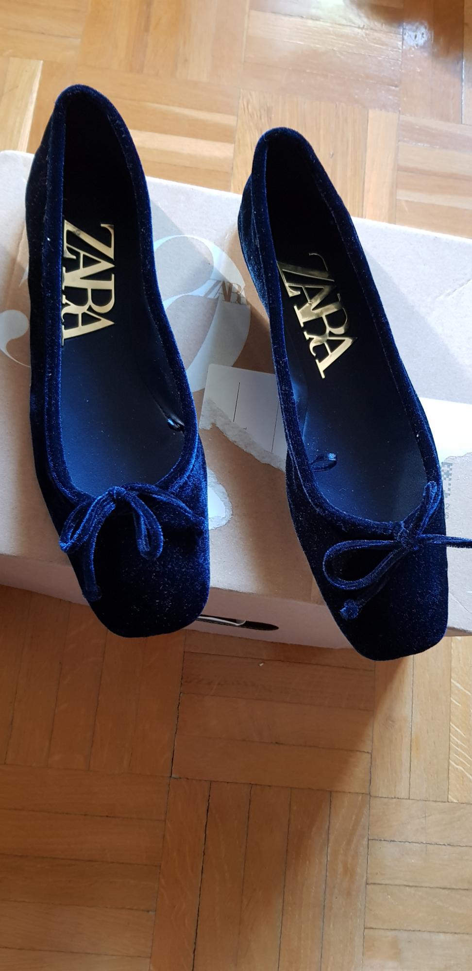 Dark blue velvet shoes with a bow, on a cardboard box. Interior with "ZARA" logo in gold letters. Wooden floor.