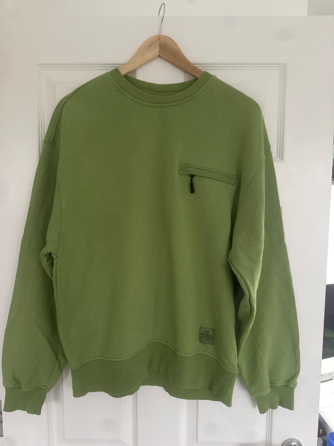 Green sweatshirt hanging on a wooden hanger over a white door. Zipped pocket on the chest and "The Original Street" label.