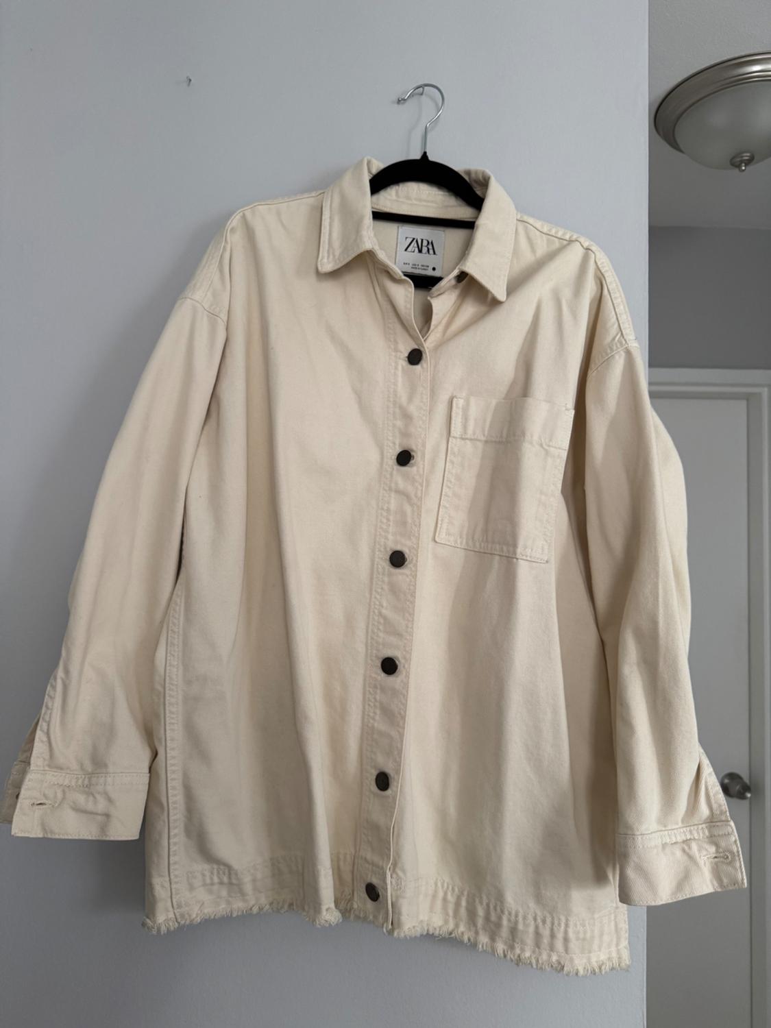 Beige long-sleeved shirt with dark buttons and a chest pocket, hanging on a black hanger. Visible label: ZARA.