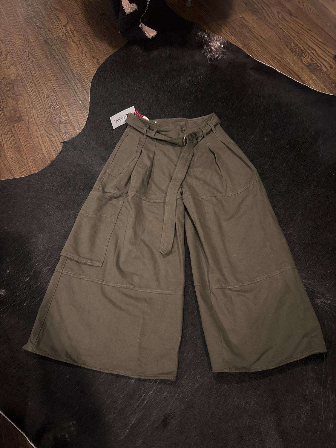 Khaki wide-leg trousers with a belt, laid on a black rug on a wooden floor. Label visible.