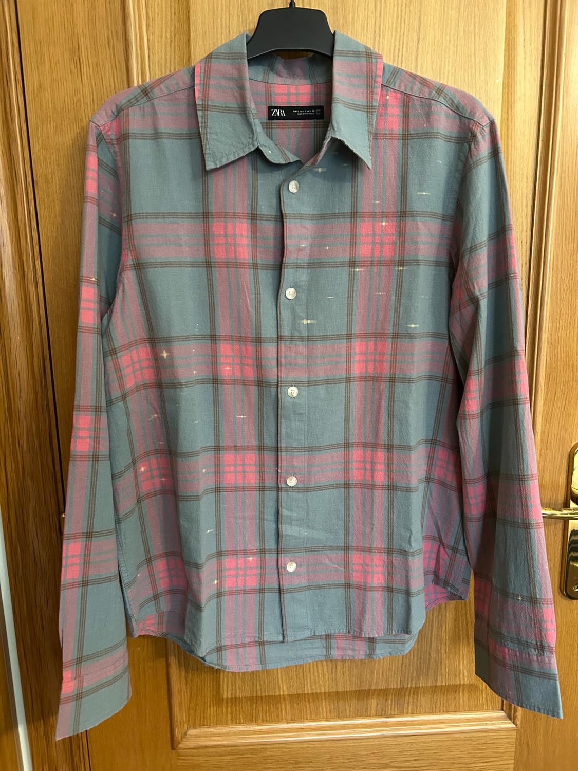 Grey and pink checked long-sleeved shirt with small spots, hanging on a hanger over a wooden door. ZARA label.