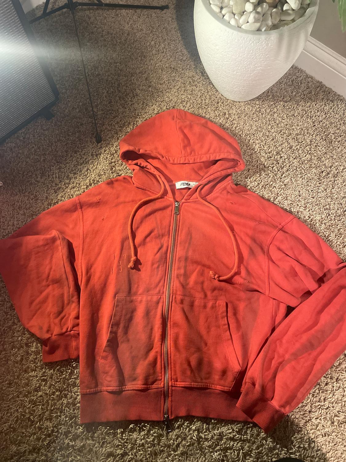 Red hooded sweatshirt with a zip on a beige rug, next to a white plant pot with decorative stones.