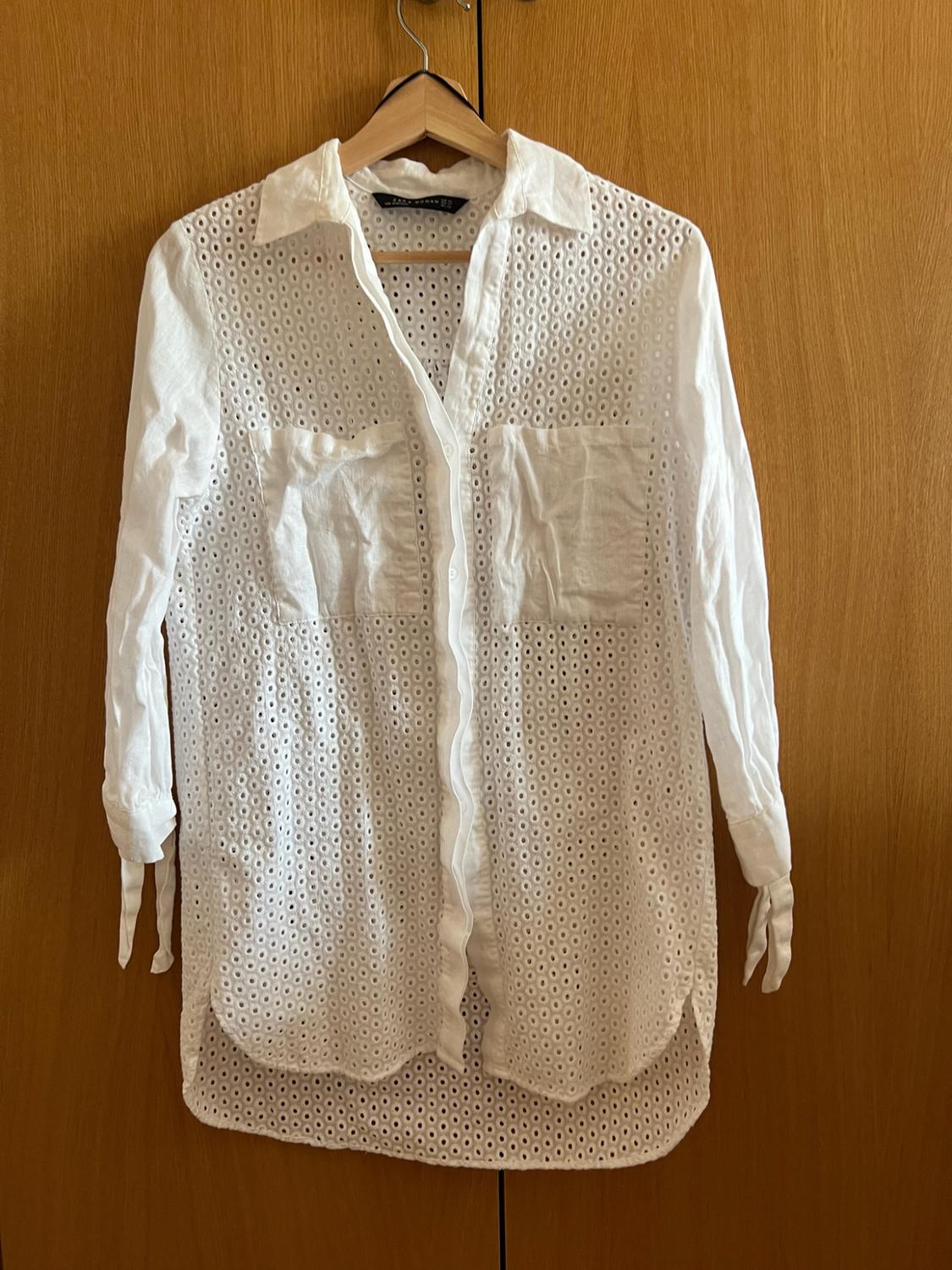 White shirt with a broderie anglaise body, pockets, and plain sleeves with bows on the cuffs, hanging against a wooden background. Text: ZARA WOMAN.