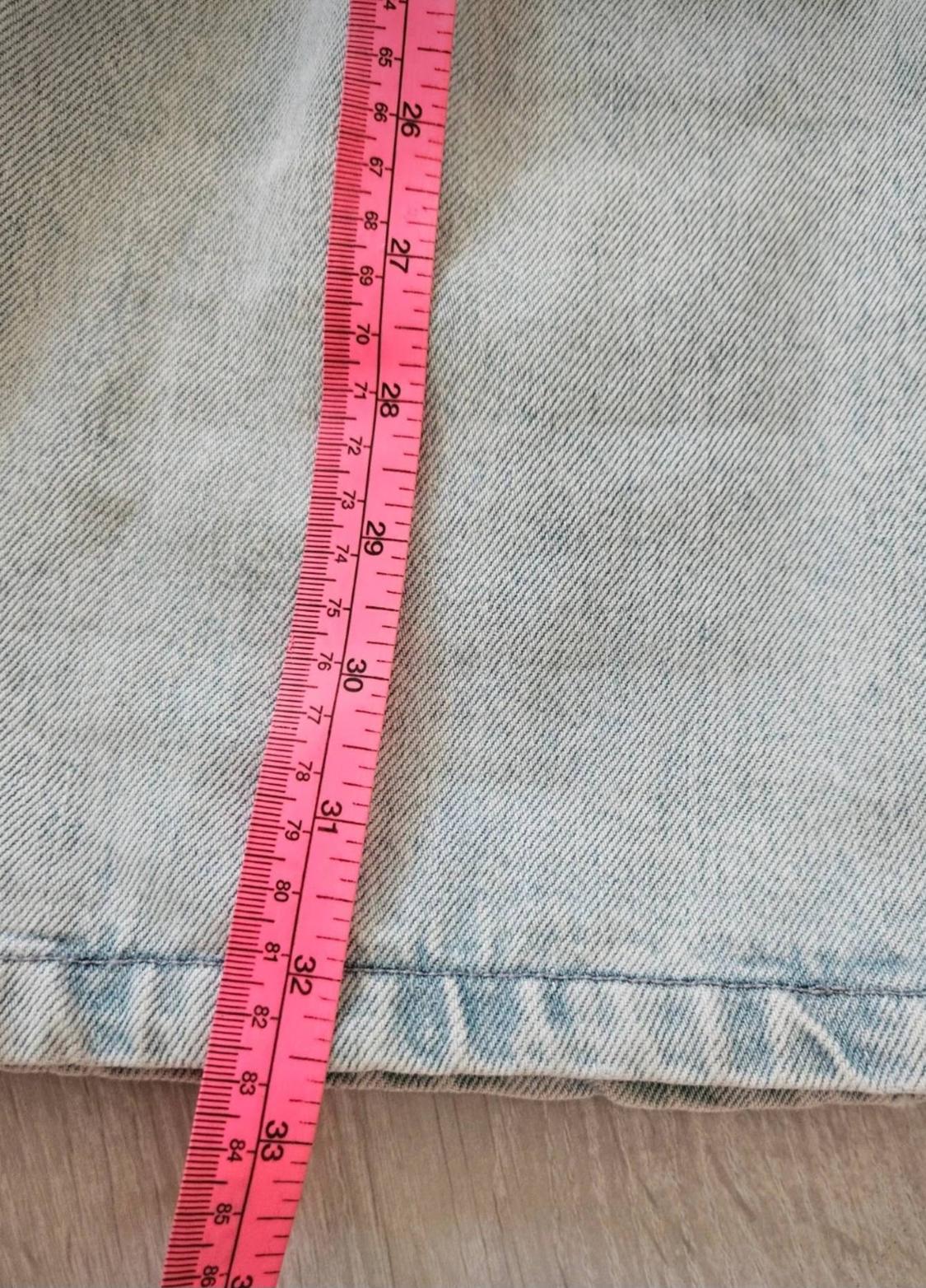 Detail of light blue denim fabric. A vertical pink tape measure on top shows the numbers 26, 27, 28, 29, 30, 31, 32 and 33.
