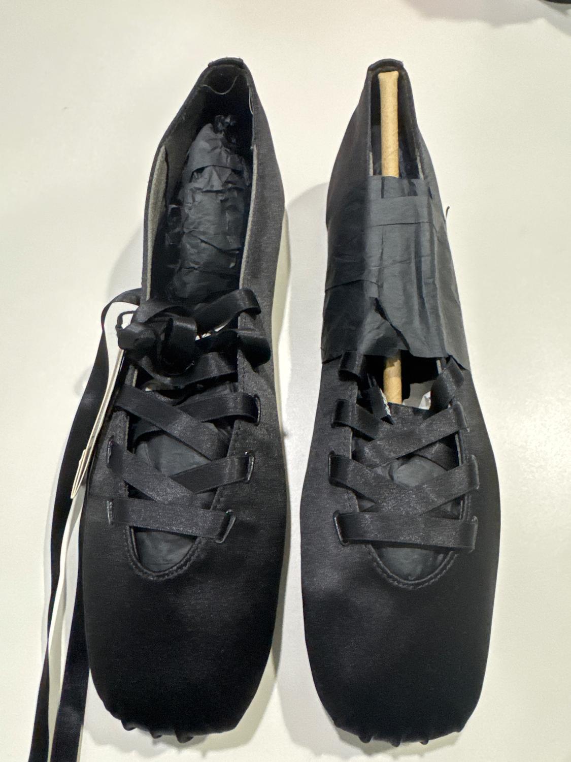 A pair of black ballet shoes on a white surface. One has a wooden stand and both have black ribbons.