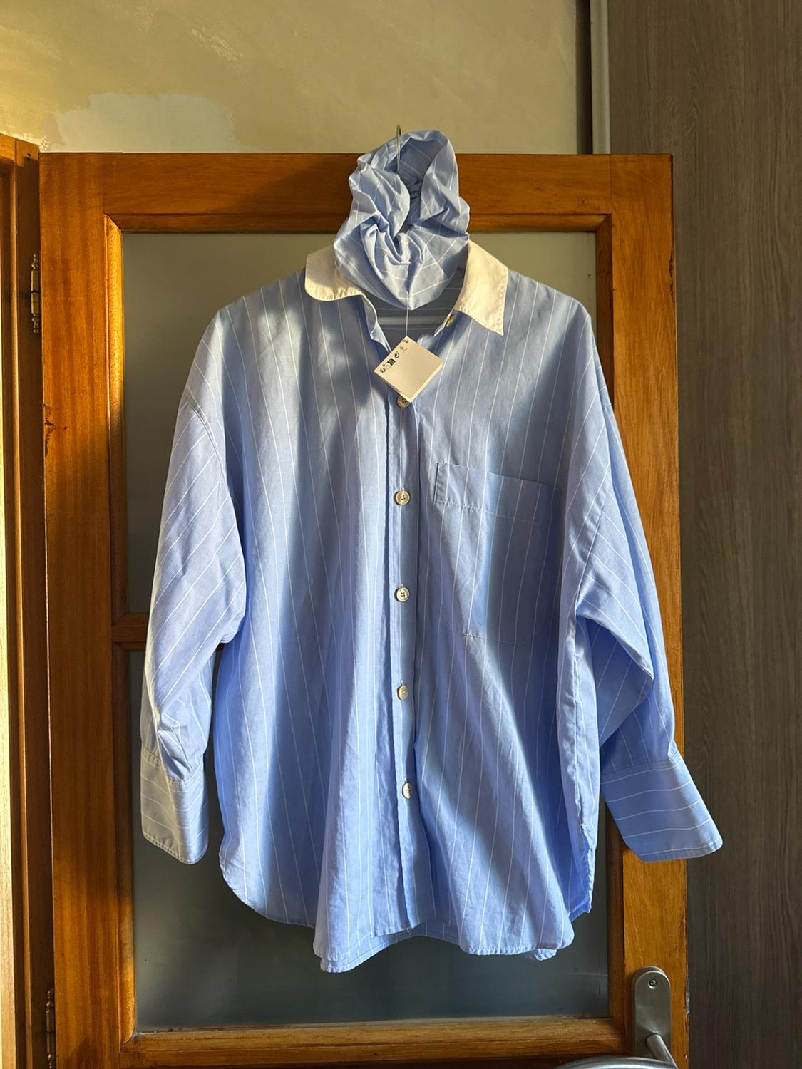 Oversized blue and white striped shirt with a white collar and matching scrunchie, hanging on a glazed wooden door in warm light.