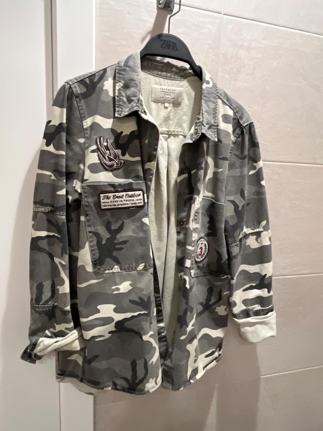 Grey camouflage jacket hanging on a black hanger with decorative patches and "The Great Outdoor" text on the chest.