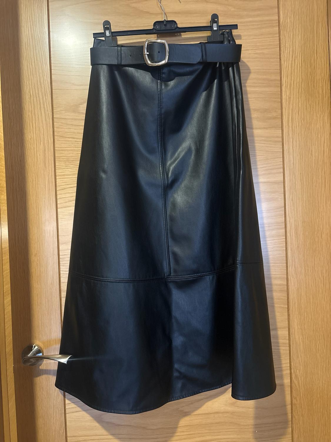 Black skirt with a belt and silver buckle, hanging on a hanger over a light wood door.