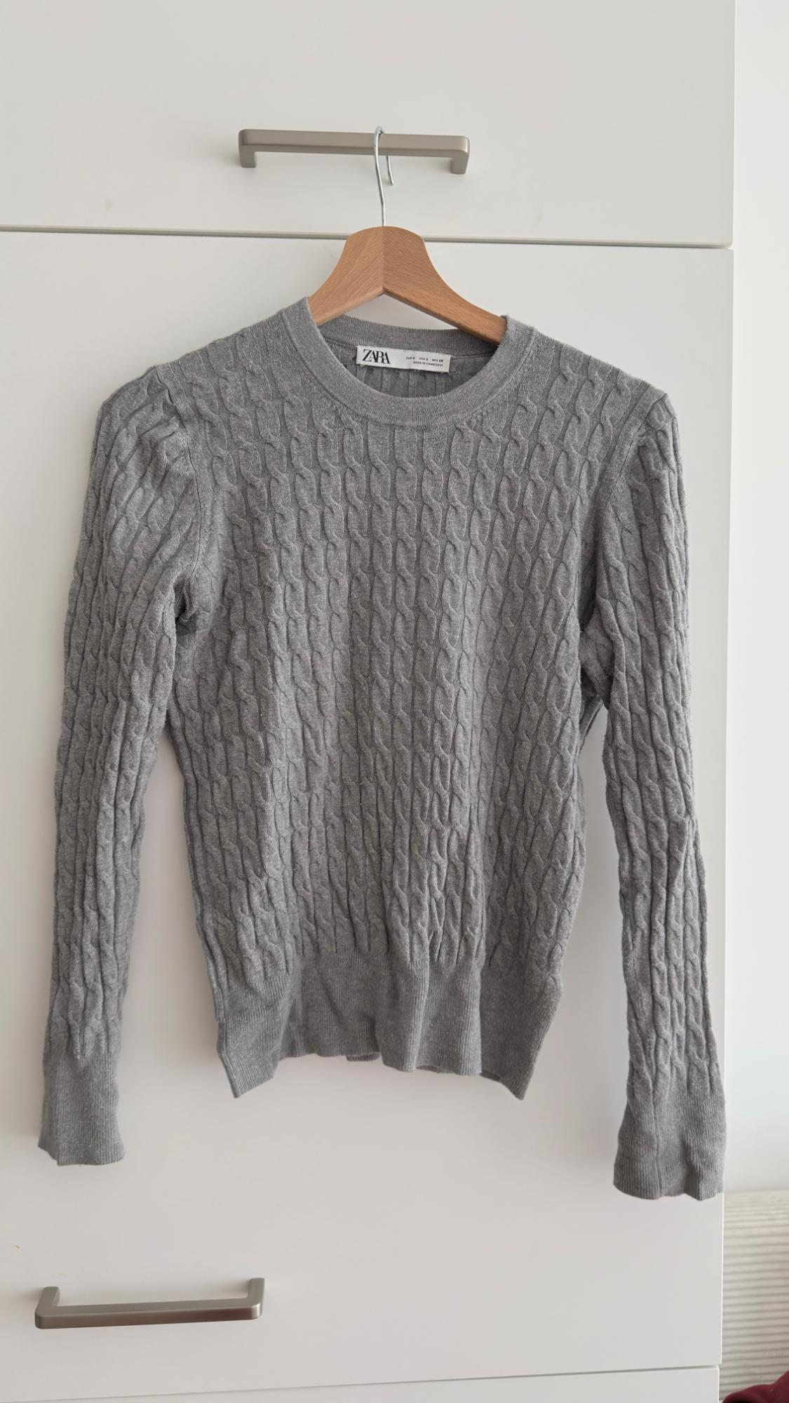 Grey cable-knit jumper hanging on a wooden hanger in front of a white wardrobe. Visible label: ZARA.