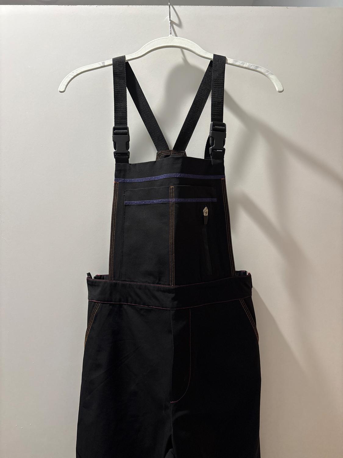 Black overalls hanging on a white hanger, with visible blue and brown stitching, against a plain white background.