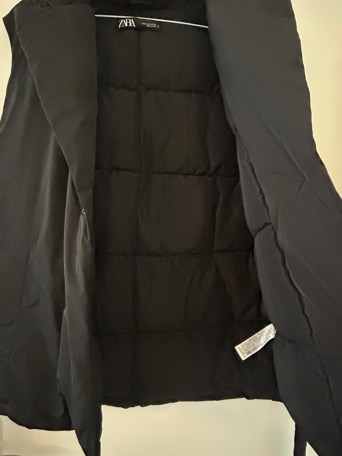 Black padded gilet hanging on a hanger, showing the inside with a visible label. Neutral setting and light background.