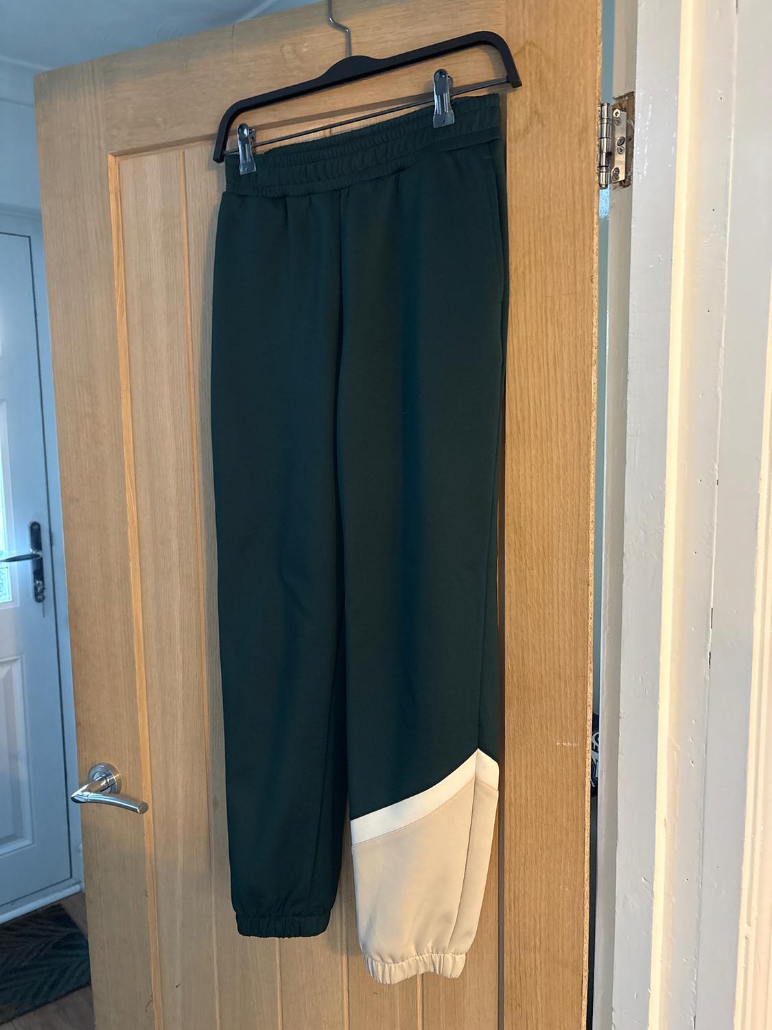 Dark green track trousers with white ankle detail, hanging on a wooden door with a black hanger.