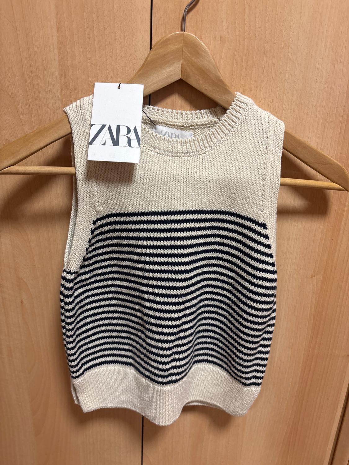 Cream-coloured sleeveless knitted top with dark horizontal stripes, on a hanger against a wooden background. Label with the text ZARA.