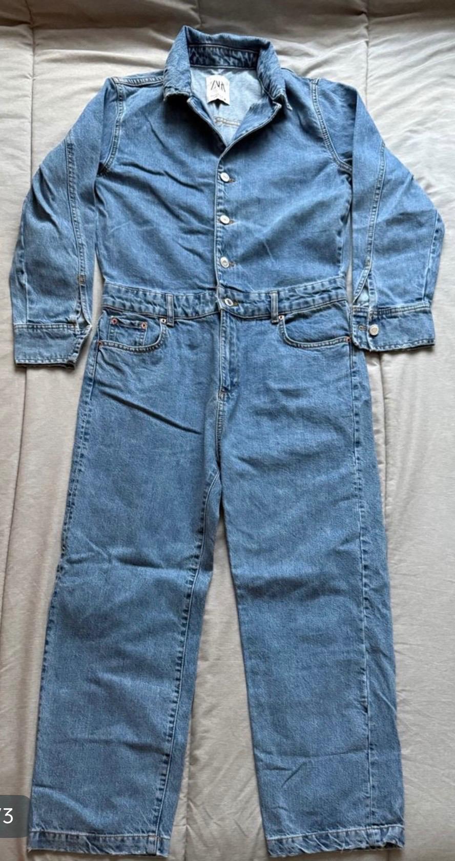 Blue denim jumpsuit with front buttons and folded collar, spread out on a padded beige surface. Visible label on the collar.