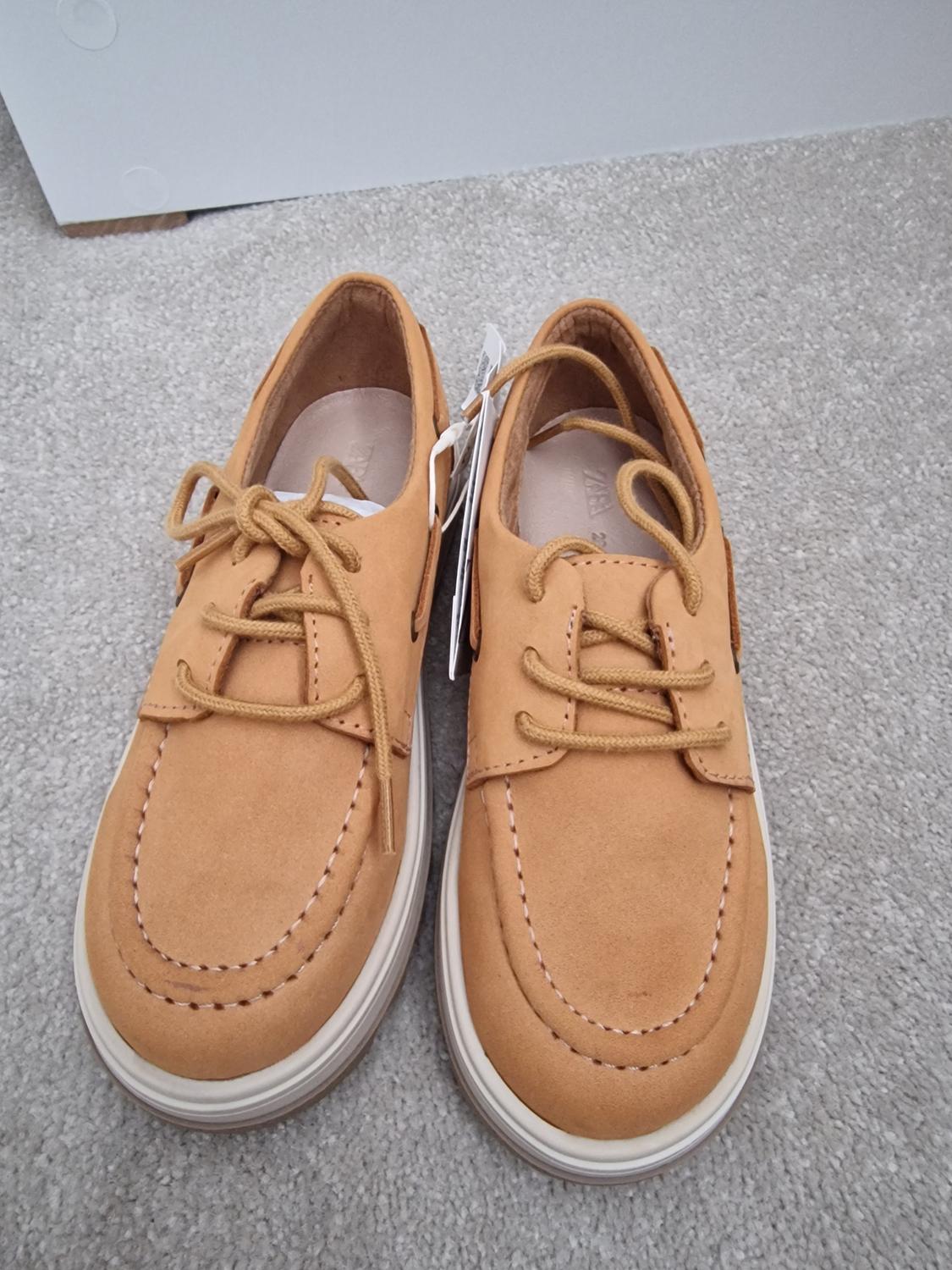 Beige suede shoes with laces, on a grey carpet. Tags hanging from one of the shoes.