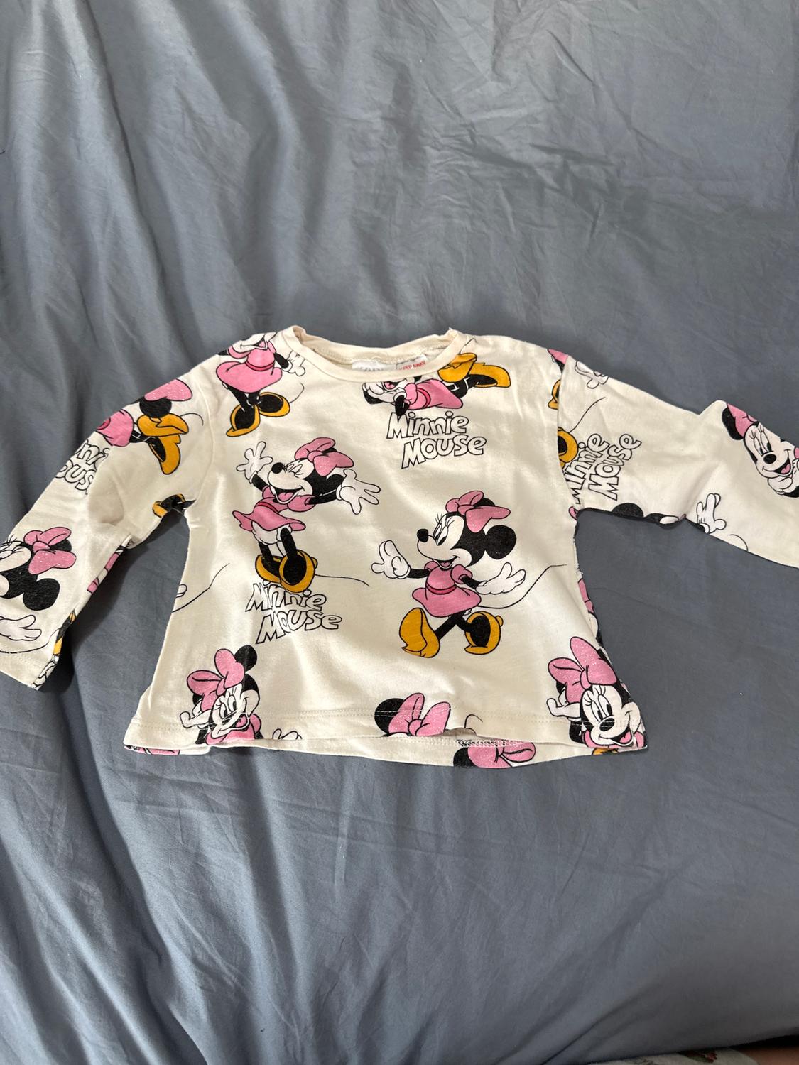 Long-sleeved children's T-shirt with a Minnie Mouse print on a white background, on a bed with grey sheets.
