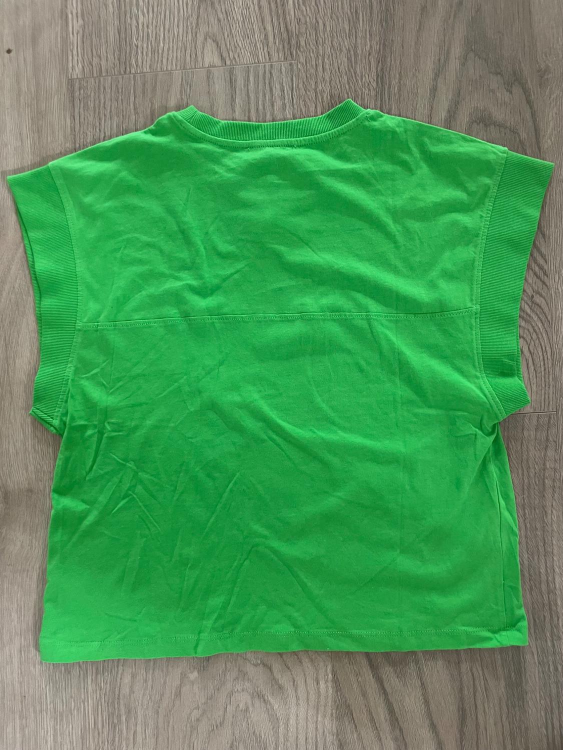 Green crew neck t-shirt with ribbed armholes and a horizontal seam on the back, laid out on a wooden floor.