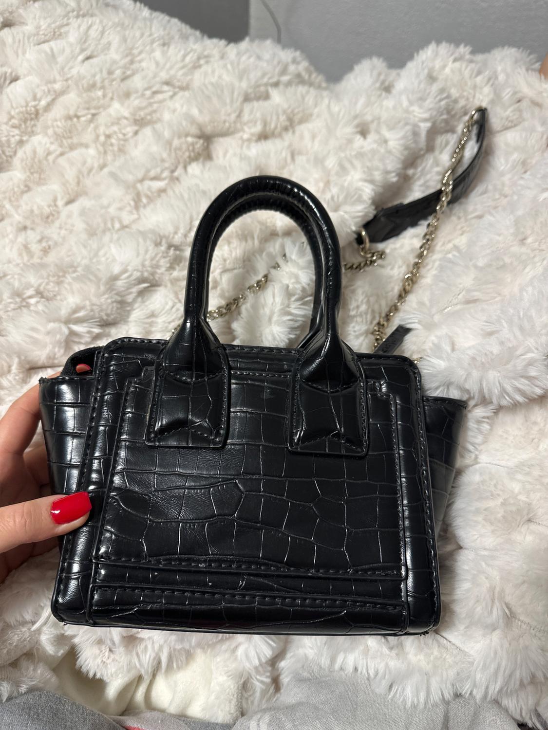 Black handbag with crocodile texture and gold chain on a white blanket. Hand with red nails holds the handbag.