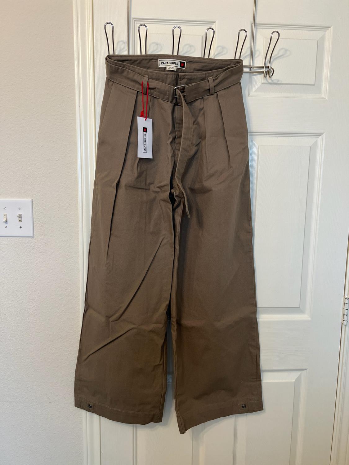 Wide brown trousers with a belt and buttons at the hem, hanging on a white door. Visible text: ZARA SRPLS.