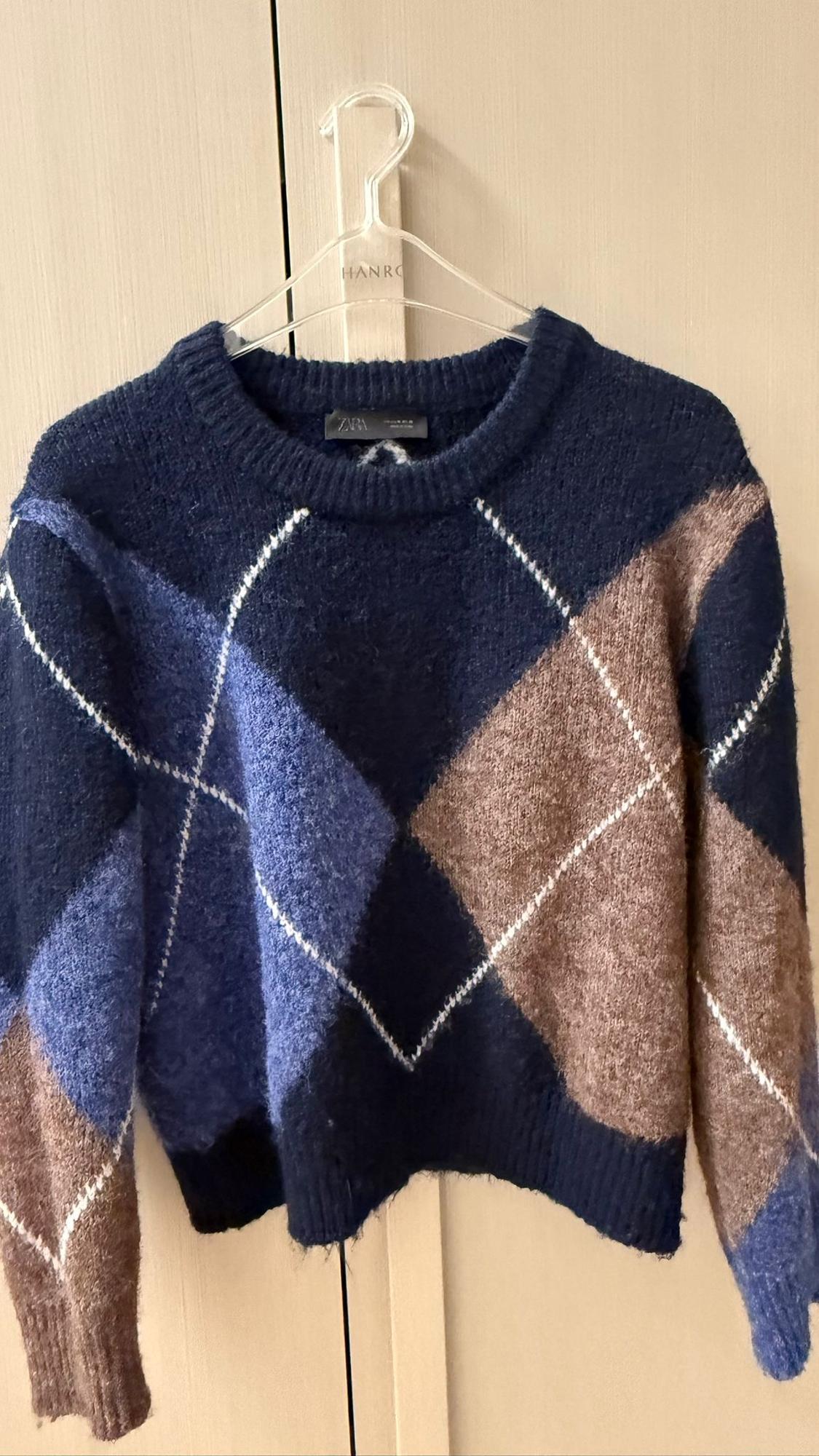 Blue and brown checked jumper hanging on a clear hanger with the brand "HANRO", against a light wooden background.