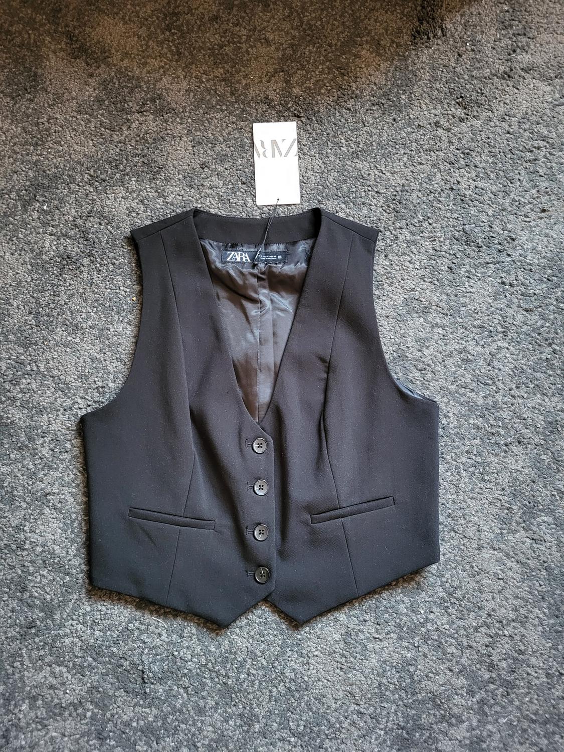 Black button-up waistcoat on grey carpet, visible label with "ZARA" text.