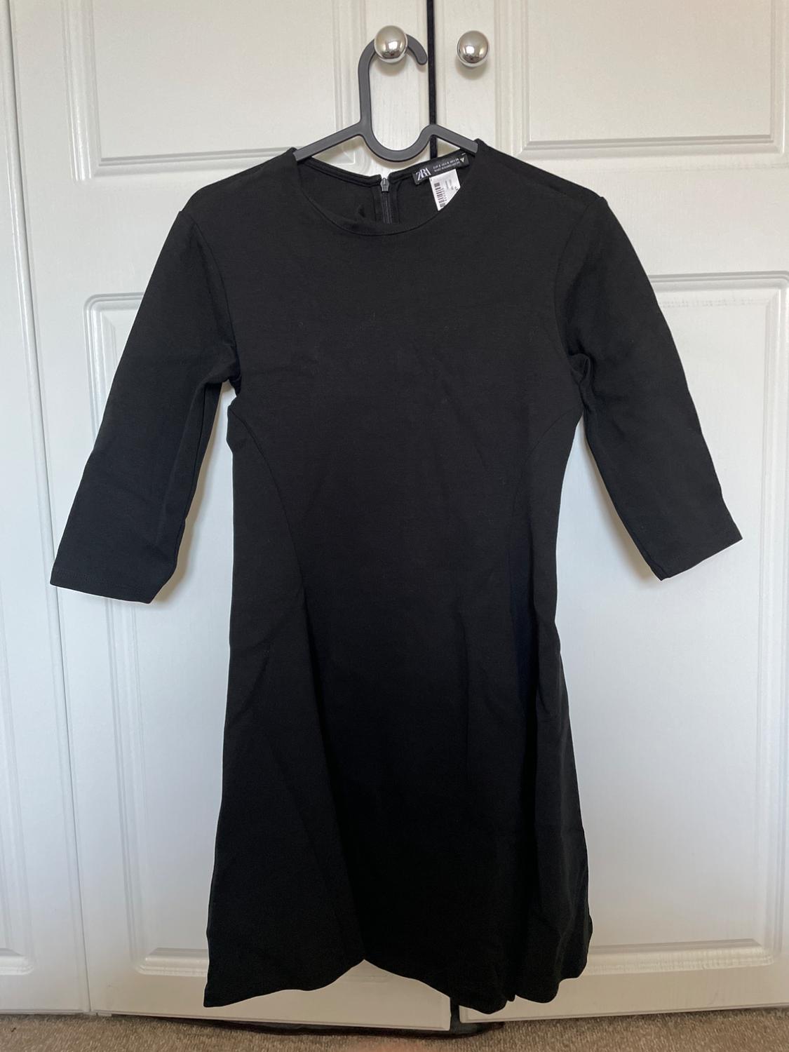 Black three-quarter sleeve, round-neck dress, hanging on a black hanger over a white door. Label with the text ZARA.