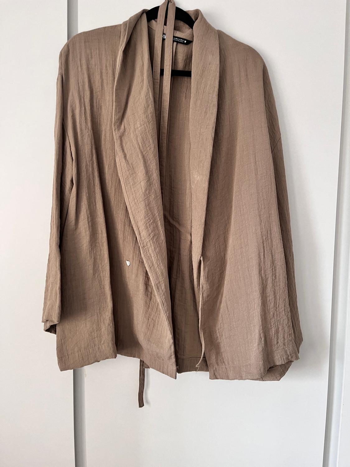 Beige lightweight fabric jacket hanging on a black hanger against a white background. Loose design with a belt in the same material.