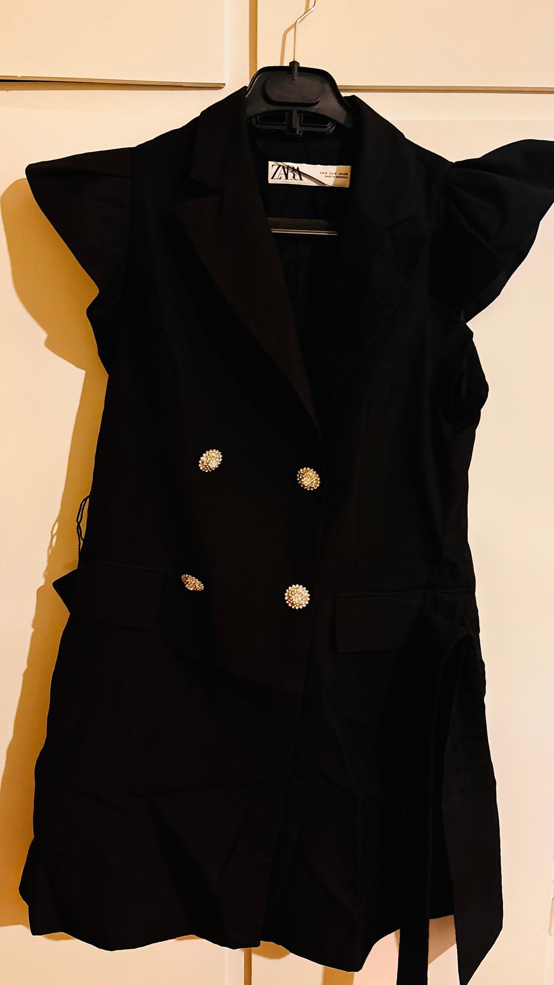 Black sleeveless waistcoat with lapels and shiny decorative buttons, hanging on a black hanger in front of a light-coloured wall.