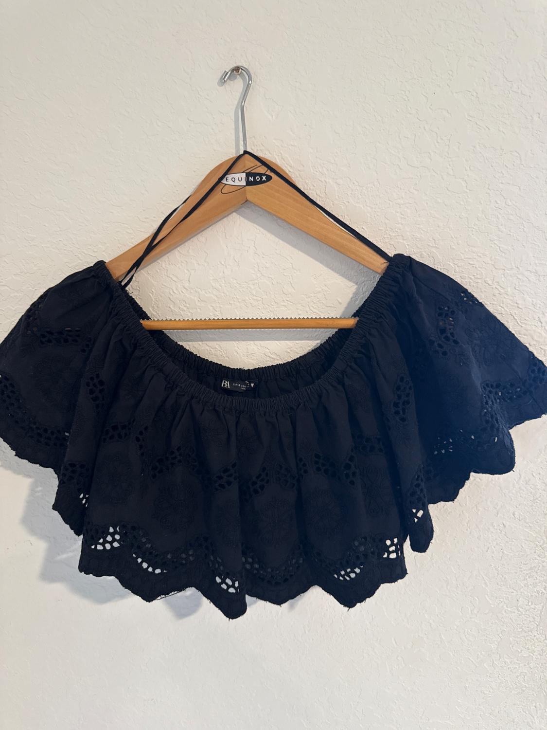 Black blouse with openwork embroidery, hanging on a wooden hanger with the "EQUINOX" brand on the white wall.
