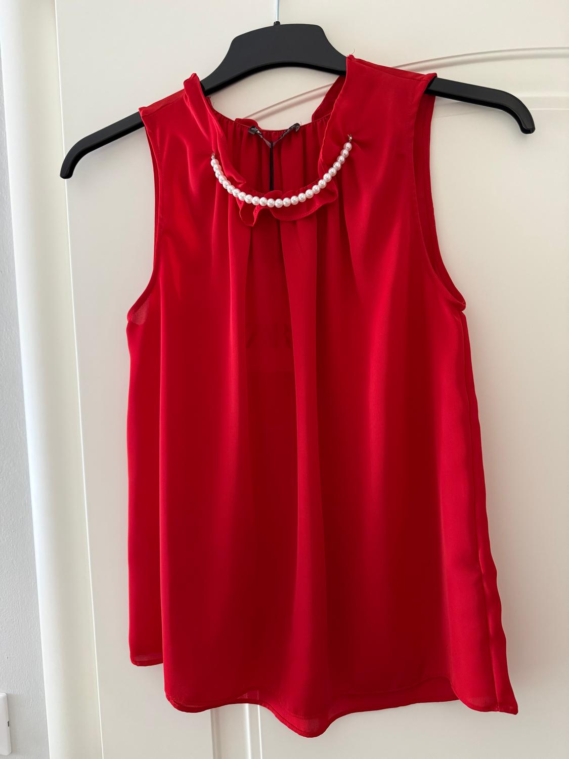 Red sleeveless blouse hanging on a black hanger, with pearl detailing on the collar, against a white background.